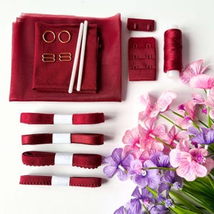 May include: A collection of burgundy lingerie-making supplies, including fabric, ribbons, hooks, rings, and thread. Two white boning rods and a spool of thread are also visible. The items are arranged on a white surface, with pink and purple flowers in the corner.