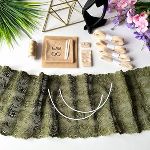 May include: An overhead shot of sewing supplies, including olive green lace, beige ribbons, thread, and various fasteners. The lace has a detailed floral pattern. Other items include a gold geometric shape and artificial flowers.