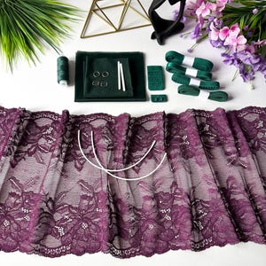 Könnte beinhalten: Eine Sammlung von Dessous-Herstellungszubehör, darunter eine breite lila Blumen-Spitze, grünes Band, Faden, BH-Zubehör und ein schwarzes Stirnband. Die Artikel sind auf einer weißen Oberfläche mit dekorativen Blumen und Grün angeordnet.
