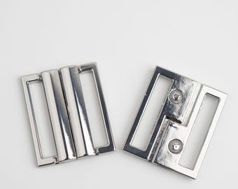 May include: Two silver metal belt buckles. One buckle has a rectangular shape with three vertical bars, and the other has a square shape with two circular rivets.