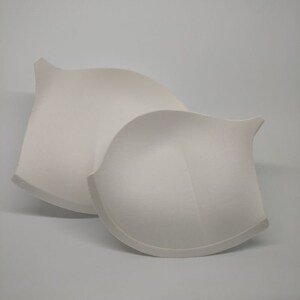 May include: Two off-white bra cups are shown against a plain backdrop. The cups have a curved form with a pointed extension at the top. The material seems to be a soft, padded fabric, indicating a focus on comfort and support.