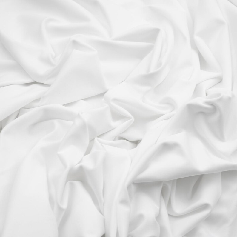 May include: A close-up shot of white fabric with soft folds and shadows. The fabric appears to be smooth and flowing, with a subtle sheen. The overall impression is one of elegance and simplicity.