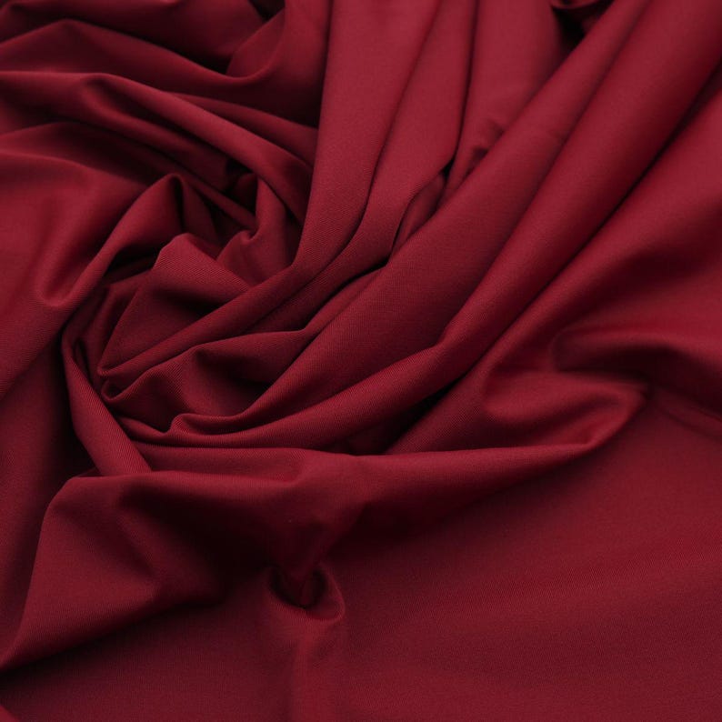 May include: Close-up of a deep red fabric with soft folds and a smooth texture. The fabric is draped, creating shadows and highlights that emphasize its flowing nature. The color is a rich, saturated shade, suggesting a luxurious material.