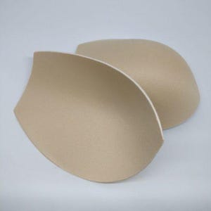 Puede incluir: Dos almohadillas beige para sujetador, diseñadas con una forma curva. Las almohadillas están hechas de un material suave similar a la espuma. La imagen está tomada sobre un fondo blanco liso, resaltando la forma y el color de las almohadillas.