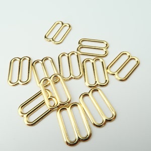 May include: A group of ten gold-toned metal slider buckles. The buckles are all the same size and shape, with two loops on each side.