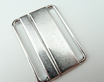 May include: A silver-colored metal bra buckle with a textured surface. The buckle has a rectangular shape with two parallel bars and two loops on each side. The background is white.
