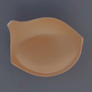May include: A beige foam bra cup insert. The insert is shaped like a half-circle and has a smooth surface.
