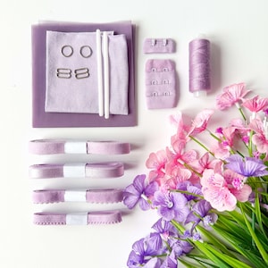 May include: A selection of lavender-coloured sewing materials, including fabric, ribbons, thread, and hook and eye closures. Silver metal rings and clasps are also present. The arrangement is complemented by a bouquet of pink and purple flowers, creating a craft-themed composition.