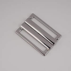 Op de afbeelding: Zilveren metalen gesp met twee rechthoekige openingen. De gesp is ontworpen om banden of riemen aan te bevestigen.
