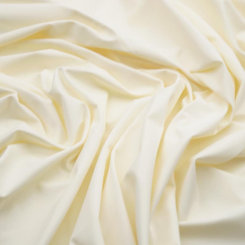 May include: A close-up shot of a cream-colored fabric with soft folds and subtle shadows. The fabric appears smooth and drapes naturally, creating a sense of movement and texture. The overall tone is neutral and elegant, suggesting a high-quality textile.