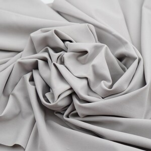 May include: Close-up of a light gray fabric with soft folds and a smooth texture. The fabric appears to be a solid color, with no visible patterns or designs. The material is likely used for clothing or home decor.