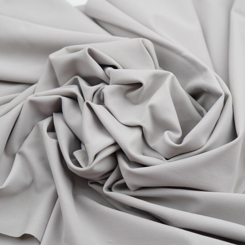 May include: Close-up of a light gray fabric with soft folds and a smooth texture. The fabric appears to be a solid color, with no visible patterns or designs. The material is likely used for clothing or home decor.