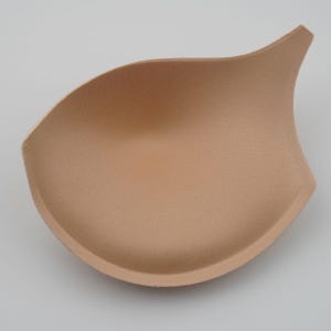Puede incluir: Un inserto de copa de sujetador de espuma beige. El inserto tiene forma de semicírculo con una punta.