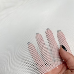 Può includere: Primo piano di tessuto bianco trasparente con un motivo a rete fine. Il tessuto è tenuto in mano, mostrando la sua trasparenza e consistenza. Il tessuto è di colore bianco brillante.