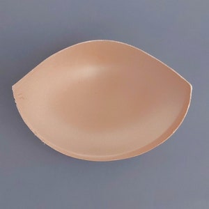 Könnte beinhalten: Ein beiger Schaumstoff-BH-Einsatz. Der Einsatz hat die Form eines Halbkreises und eine glatte Oberfläche.