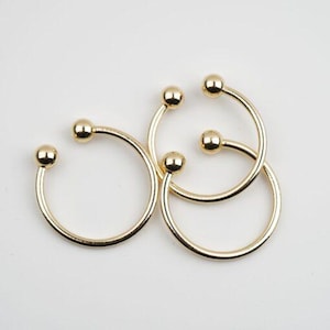 Könnte beinhalten: Drei goldfarbene Metallringe mit abgerundeten Kanten und kugelförmigen Enden. Die Ringe sind ineinander verschlungen.