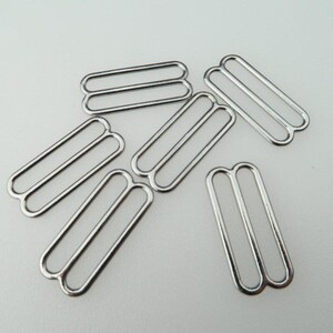 May include: A collection of six silver-colored metal strap adjusters, each featuring a rectangular frame with a central bar. These hardware components are designed with rounded corners and a smooth, reflective finish, arranged in a scattered pattern.