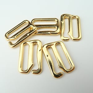 May include: Several gold-colored metal bra strap hooks and sliders are scattered on a white surface. The hooks have a curved design, and the sliders are rectangular. These components are used in lingerie and garment construction.