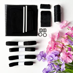 Puede incluir: Un conjunto de suministros de costura negros, que incluyen tela, cinta, hilo y componentes para hacer sujetadores, dispuestos sobre una superficie blanca. Dos varillas blancas y un ramo de flores artificiales rosas y moradas completan la composición.