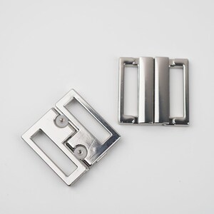 May include: Two silver metal bra strap buckles. One is open and the other is closed. The buckles have a rectangular shape with rounded corners and a shiny finish. The background is white.
