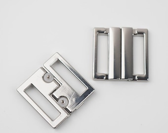 May include: Two silver metal bra strap buckles. One is open and the other is closed. The buckles have a rectangular shape with rounded corners and a shiny finish. The background is plain white.