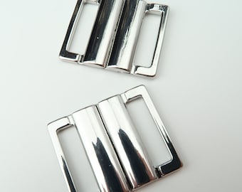 May include: Two silver metal clasps with a double-bar design. The clasps are commonly used for swimwear or lingerie.