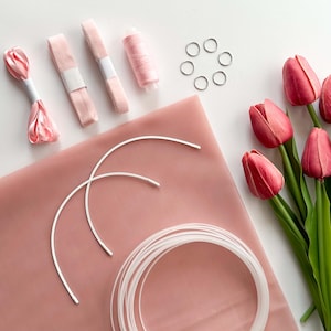May include: A flat lay featuring craft supplies in shades of pink and white. Items include ribbon, thread, metal rings, and curved wire. Fresh tulips with pink petals and green stems are arranged on the right side of the image.