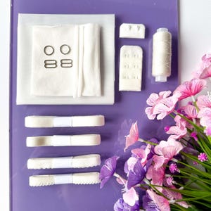 May include: A craft kit with white fabric, metal rings, bra hooks, and spools of thread. The kit includes several rolls of white ribbon and is arranged on a purple surface. Pink and purple flowers are in the background.