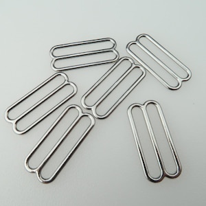 May include: A set of six silver metal slider buckles. The buckles are rectangular in shape with two parallel loops. They are commonly used for adjusting the length of straps or belts.