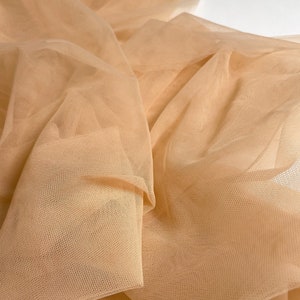 May include: A close-up of a piece of beige tulle fabric. The fabric is soft and delicate, with a fine mesh weave.