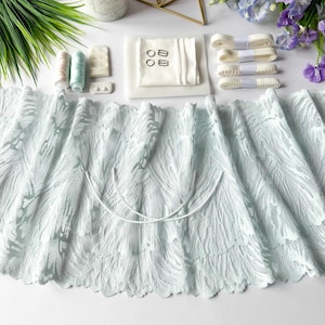 May include: A pale blue lace fabric with a feather design is the central element. White ribbons, thread spools, and sewing supplies are arranged around the lace. The image suggests a craft or sewing project, with a focus on textiles.
