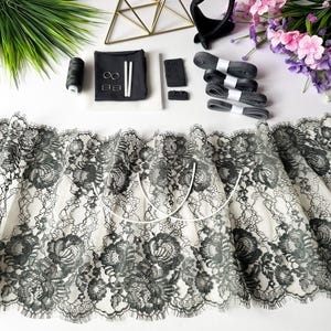 May include: A collection of lingerie-making supplies, including black lace fabric with a floral pattern, gray ribbons, and various metal and plastic hardware. A spool of black thread, white boning, and a black fabric pouch are also present.