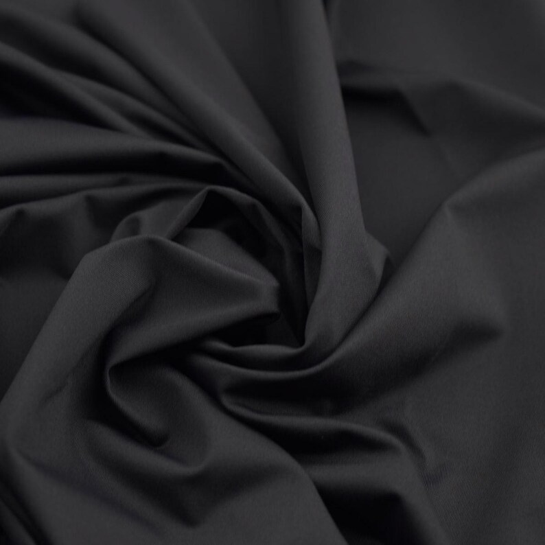 May include: A close-up shot of a dark gray fabric with a smooth texture. The fabric is draped and swirled, creating soft folds and shadows. The material appears to be a solid color, with no visible patterns or designs.