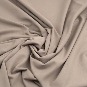 May include: Close-up of a beige fabric with soft folds and a textured surface. The fabric is tightly woven and appears to be a knit or jersey material, suitable for clothing or home decor projects.