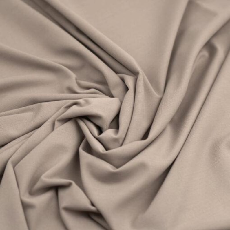 May include: Close-up of a beige fabric with soft folds and a textured surface. The fabric is tightly woven and appears to be a knit or jersey material, suitable for clothing or home decor projects.