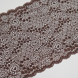 Op de afbeelding: Close-up van een gedeelte van bruine kanten stof. Het kant heeft een delicaat, open ontwerp met een geschulpte rand. De kleur is een rijk, donkerbruin en de textuur lijkt zacht en ingewikkeld. Het kant wordt waarschijnlijk gebruikt voor kleding of knutselprojecten.
