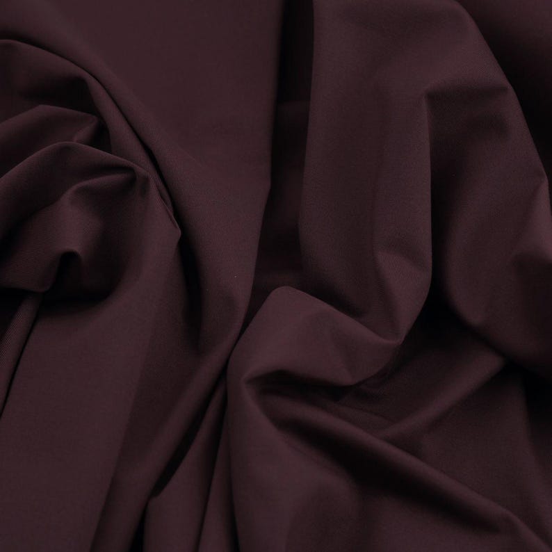 May include: A close-up shot of a deep burgundy fabric with soft folds and shadows. The material appears to be a smooth, solid color, possibly cotton or a similar textile. The fabric is draped, creating a sense of depth and texture.