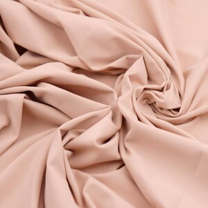 May include: Close-up of a peach-colored fabric with soft folds and a smooth texture. The fabric is draped, creating shadows and highlights that emphasize its flow and drape. The overall appearance suggests a lightweight material.