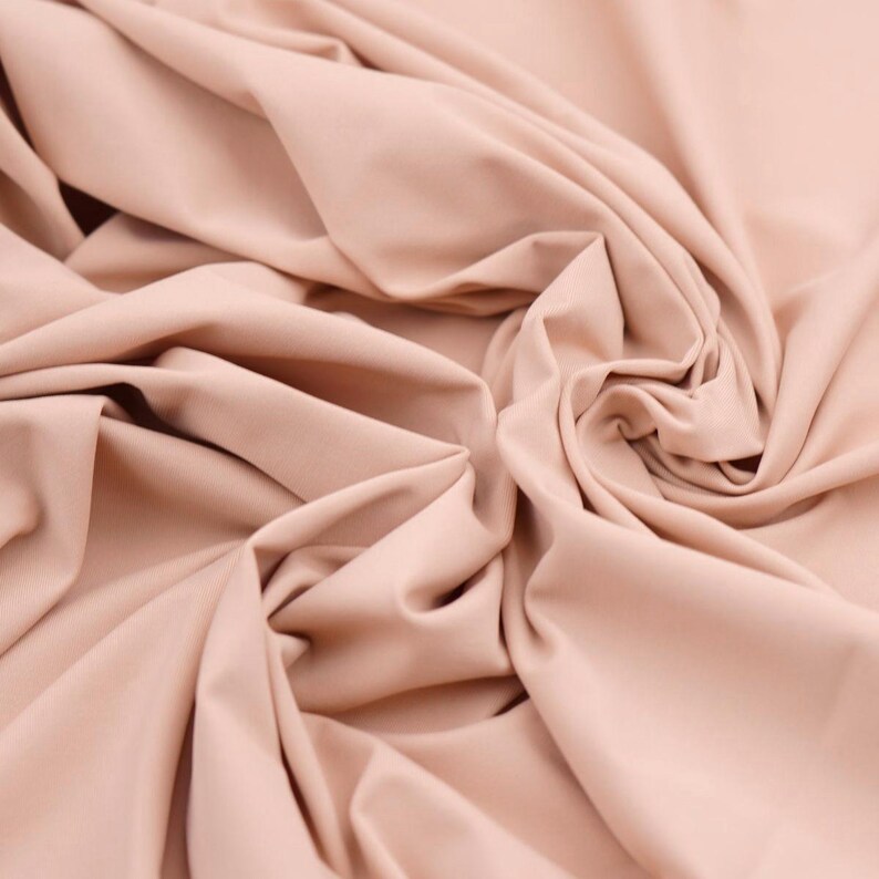 May include: Close-up of a peach-colored fabric with soft folds and a smooth texture. The fabric is draped, creating shadows and highlights that emphasize its flow and drape. The overall appearance suggests a lightweight material.