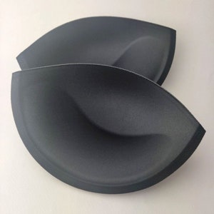 May include: Two black bra cups, each with a curved, petal-like shape. The cups are made of a textured material and are designed to provide support and shaping. The background is a plain white surface.