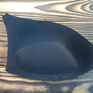 May include: Black bra cup insert with a smooth, slightly textured surface. The insert has a rounded shape and is designed to provide lift and support.