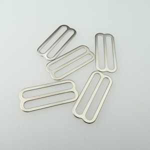 May include: A collection of five silver-colored metal bra strap sliders. Each slider has a rectangular shape with two parallel slots, designed for adjusting straps. The sliders are scattered against a plain white background.