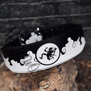 May include: Black and white leather belt with a decorative design. The belt features a circular graphic with a running figure and cloud-like patterns. The belt is approximately 5 cm wide.