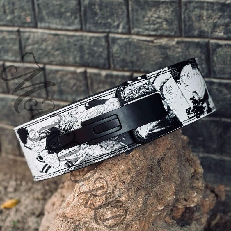 May include: A black weightlifting belt with a white and black manga-style print. The belt has a black metal buckle.