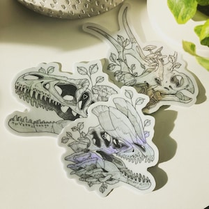 May include: Three black and white dinosaur skull stickers with floral details. The stickers are on a white background.