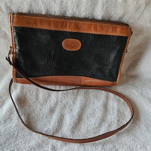 Puede incluir: Un bolso de hombro de cuero negro y marrón con una cremallera dorada y un logotipo que dice "Marco Avané".