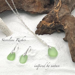 May include: A silver necklace and earrings set featuring pale green sea glass pendants. The earrings have a hook design. The necklace and earrings are displayed on a white surface with driftwood in the background. The text "Shoreline Riches... inspired by nature" is also visible.