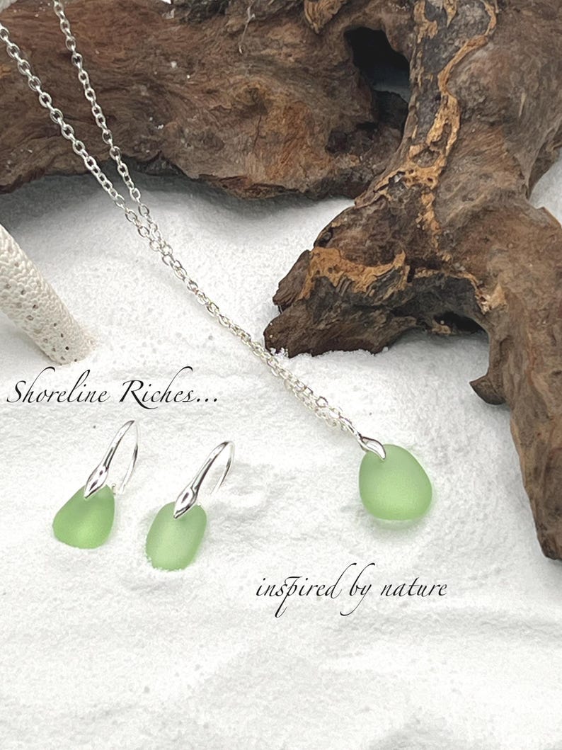 May include: A silver necklace and earrings set featuring pale green sea glass pendants. The earrings have a hook design. The necklace and earrings are displayed on a white surface with driftwood in the background. The text "Shoreline Riches... inspired by nature" is also visible.