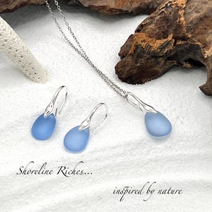 May include: A silver necklace and earrings set featuring teardrop-shaped, light blue pendants. The earrings have a hook design. The text "Shoreline Riches... inspired by nature" is also visible.
