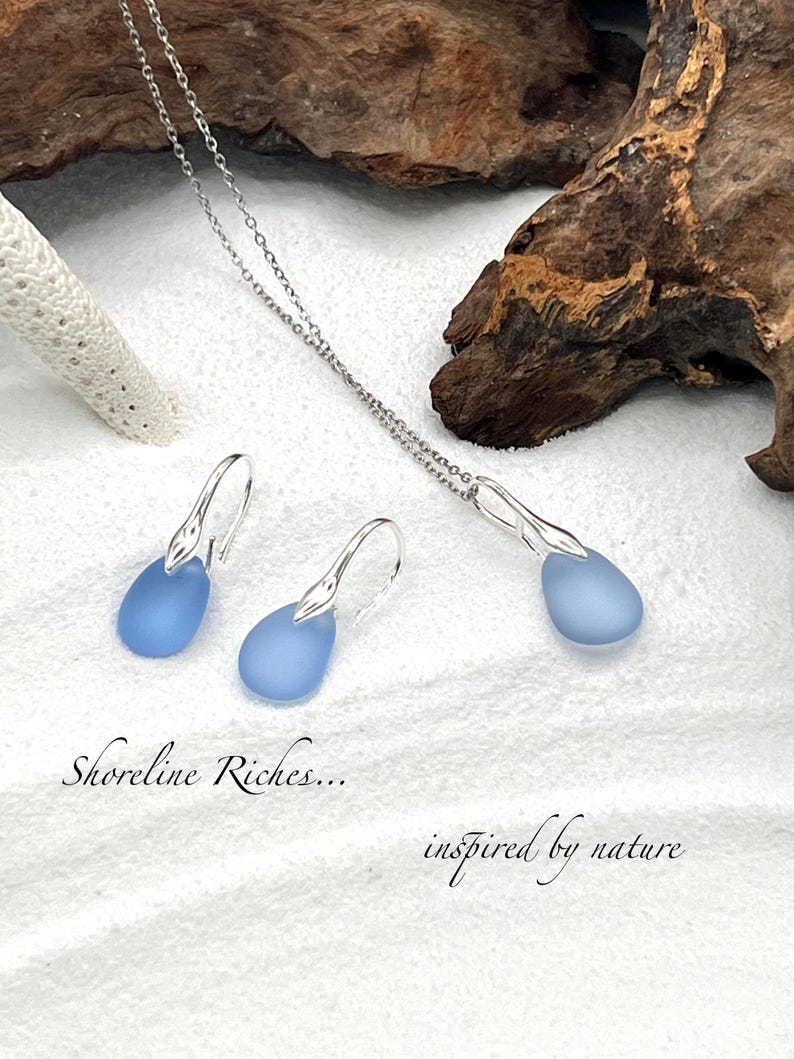 May include: A silver necklace and earrings set featuring teardrop-shaped, light blue pendants. The earrings have a hook design. The text "Shoreline Riches... inspired by nature" is also visible.